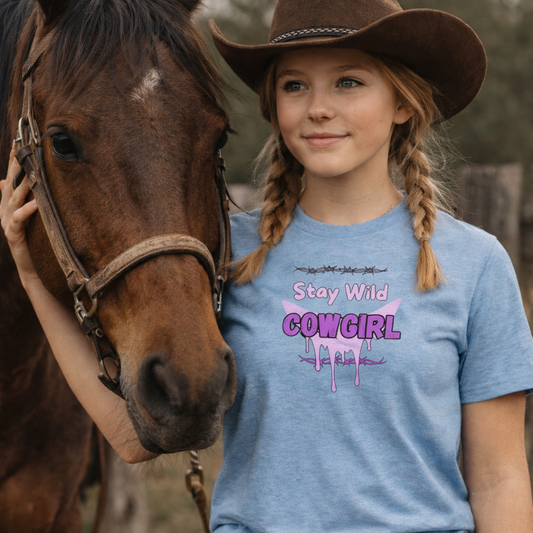 A girl wearing a Cowgirl Western Tee with 'Stay Wild COWGIRL' graphic next to a horse, showcasing a playful cowgirl vibe.
